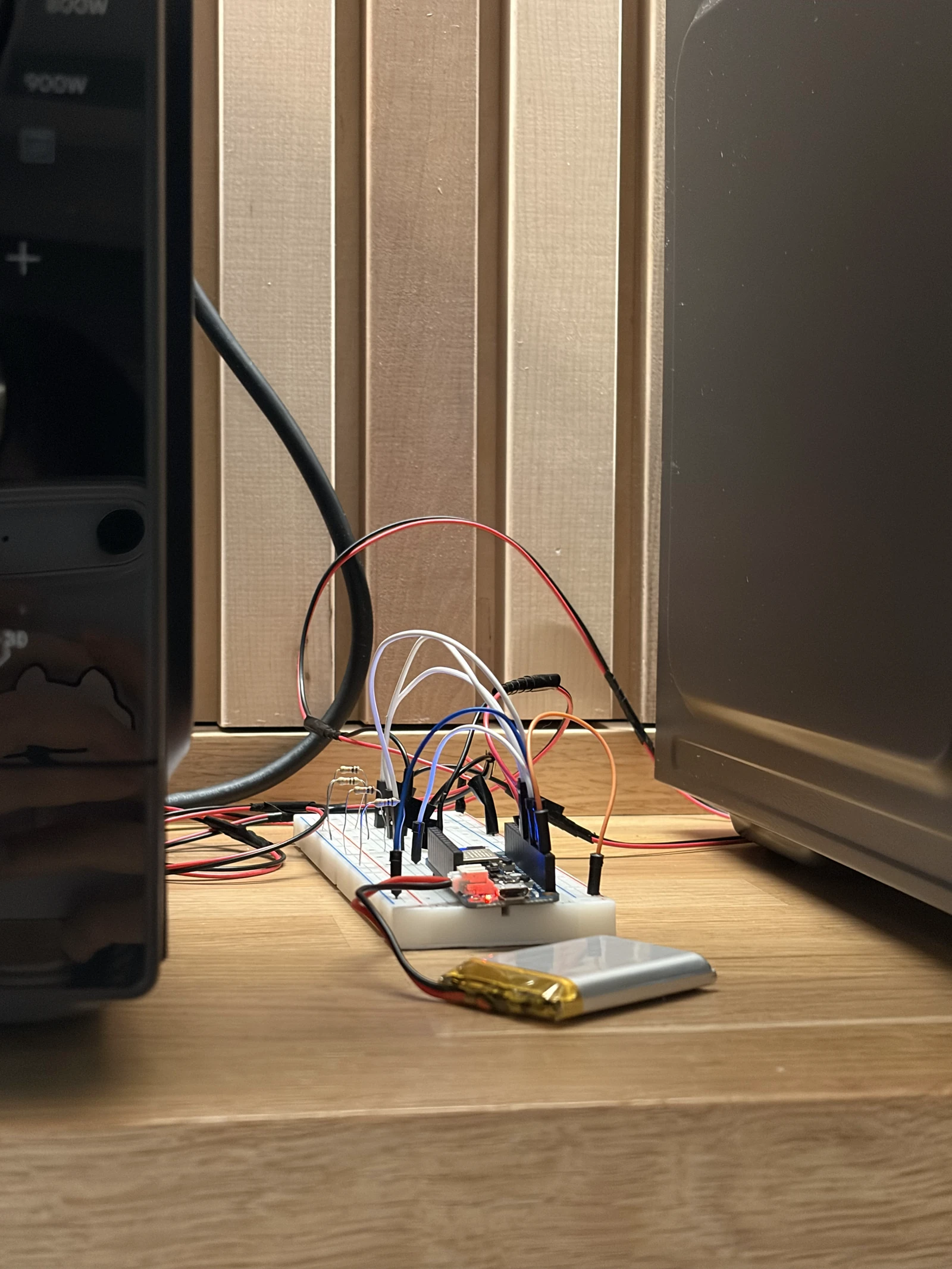 A breadboard with an Arduino Feather and a battery, wired to the photosensors, on the counter between two microwaves